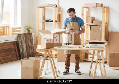 Full length portrait of mature bearded man wearing apron packaging individual food portions, worker in food delivery service, copy space Stock Photo