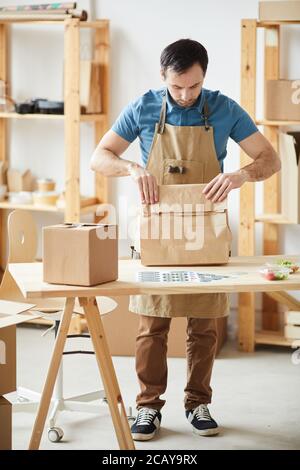 Full length portrait of mature man wearing apron packaging orders while standing by wooden table, food delivery service worker Stock Photo
