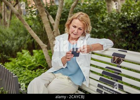 Charming caucasian senior woman about 60 years old with cell phone in hands is sitting on the bench with social distancing markup in public park. She Stock Photo