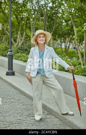 Caucasian aged woman about 60 years old is standing in public pakr in casual cloth and with orange umbrella. Elderly lady is travelling, she is active Stock Photo
