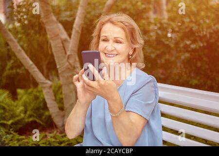 Elderly caucasian attractive lady about 62 years old is using her cell phone  while sitting on the bench in public park. She smiles and looks friendly Stock Photo