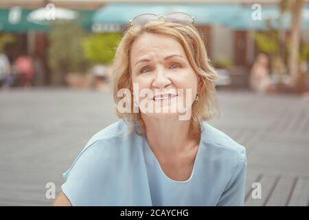 Portrait of elderly caucasian pretty woman about 62 years old in public park. She smiles and looks active, cheerful and happy lady. She wears sunglass Stock Photo