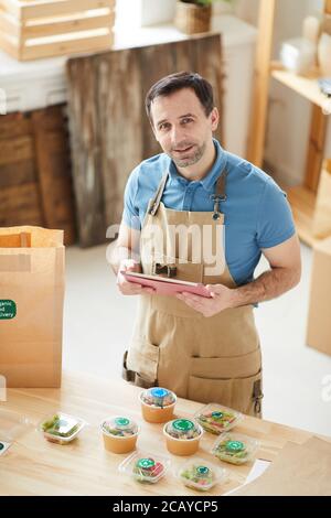 High angle portrait of mature man wearing apron looking at camera while packaging orders at wooden table in food delivery service