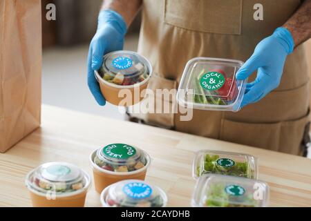 Closeup of unrecognizable worker wearing protective gloves safely packaging orders at wooden table in food delivery service, copy space Stock Photo