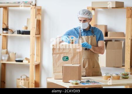 Waist up portrait of mature male worker wearing protective clothes while safely packaging orders at wooden table in food delivery service, copy space Stock Photo