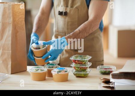 Closeup of unrecognizable worker wearing protective gloves packaging orders at wooden table in food delivery service, copy space Stock Photo