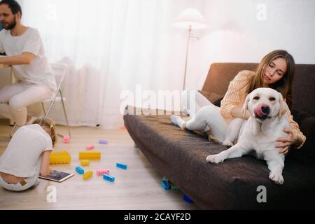 friendly family spend leisure time at home together, everyone is busy with their own interests, mother playing with pet dog, little daughter with toys Stock Photo