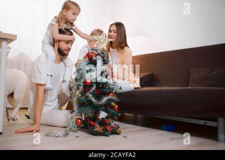 beautiful caucasian family at home, decorating christmas tree together. Cute little kid girl sit on neck of father and putting decorative star on the Stock Photo