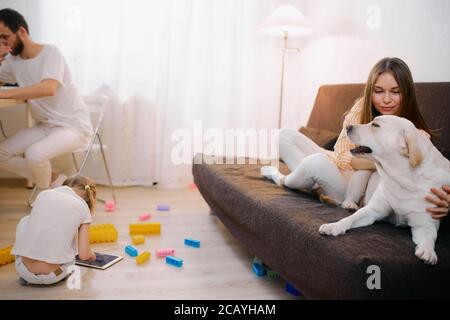 adorable family consisted of mother, father and kid girl at home in living room, in casual white clothes. children playing on the floor, while their p Stock Photo