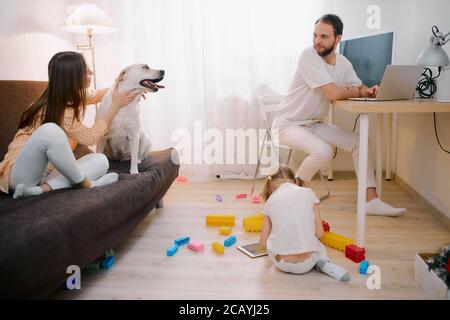 family together at home, spend free time, holiday in living room, have a rest. Child girl play with toys, woman petting their domestic dog, while man Stock Photo