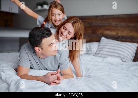 portrait of beautiful happy family lying on bed together, mother father and kid girl kissing hugging and laughing. indoors Stock Photo