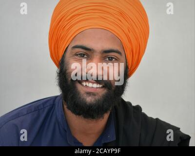 Handsome young Indian Sikh devotee with black beard and smiling eyes wears an orange turban (dastar) and looks into the camera. Stock Photo