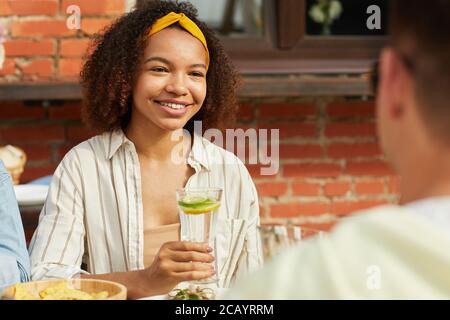 Portrait of smiling African-American woman enjoying dinner with friends outdoors and holding refreshing cocktail while sitting at table during Summer Stock Photo