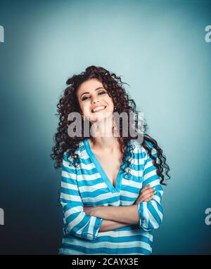 Success. Closeup portrait head shot confident beautiful happy young brunette curly woman arms crossed smiling isolated blue background wall. Positive Stock Photo