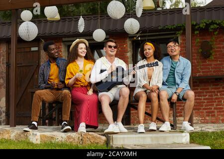 Full length portrait of young people enjoying outdoor party in Summer with focus on man playing guitar and singing in foreground, copy space Stock Photo