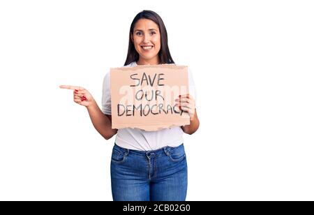 Young beautiful woman holding save our democracy cardboard banner smiling happy pointing with hand and finger to the side Stock Photo