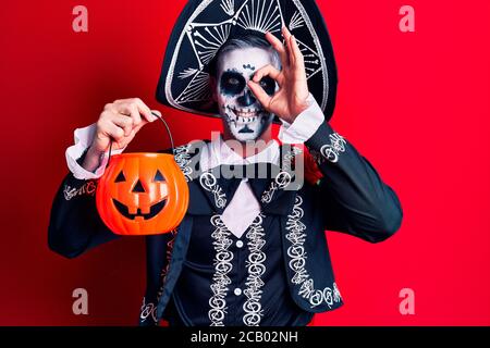Young man wearing mexican day of the dead costume holding pumpkin smiling happy doing ok sign with hand on eye looking through fingers Stock Photo