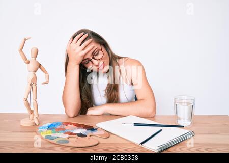 Beautiful young caucasian woman artist painter sitting on desk surprised with hand on head for mistake, remember error. forgot, bad memory concept. Stock Photo