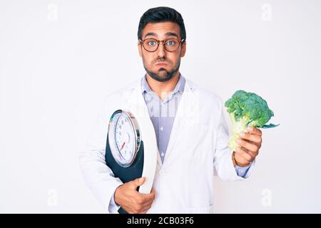 Young hispanic man as nutritionist doctor holding weighing machine and broccoli puffing cheeks with funny face. mouth inflated with air, catching air. Stock Photo