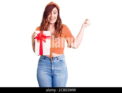 Young beautiful woman holding gift screaming proud, celebrating victory and success very excited with raised arms Stock Photo
