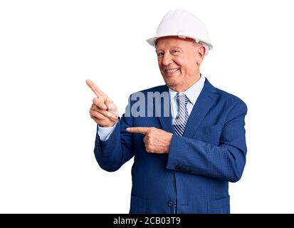 Senior handsome grey-haired man wearing suit and architect hardhat smiling and looking at the camera pointing with two hands and fingers to the side. Stock Photo