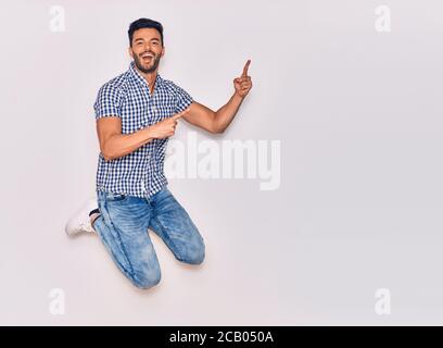 Young handsome hispanic man wearing casual clothes smiling happy. Jumping with smile on face pointing with fingers to the side over isolated white bac Stock Photo