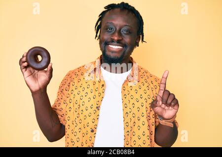 Young african american man with braids holding donut surprised with an idea or question pointing finger with happy face, number one Stock Photo