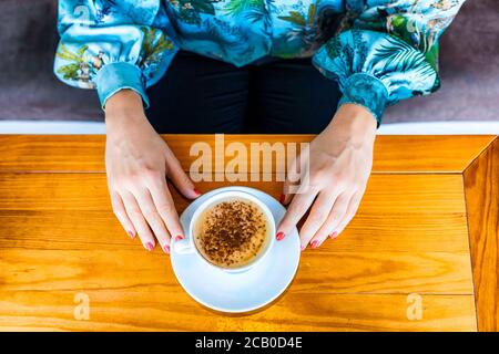 View from the top of two hands of a woman with red polish nail while drinking a cappuccino in a local coffee house in Lisbon