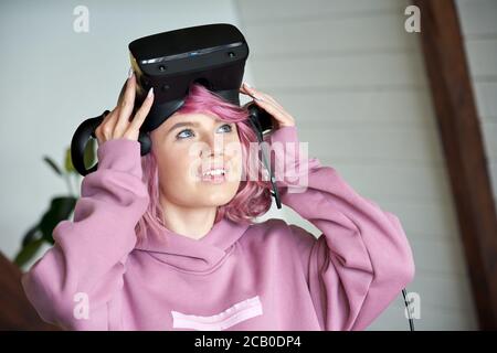 Excited teen girl with pink hair wear vr headset hold controller looking up. Stock Photo