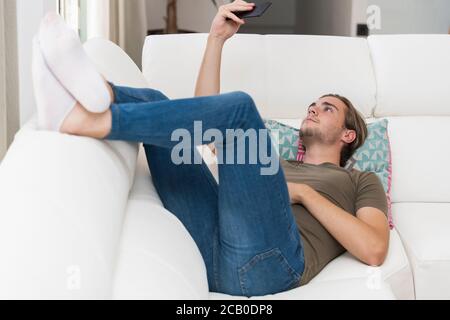Relaxed looking handsome young man lying on a sofa and watching his phone. Relaxation and technology concept. Stock Photo