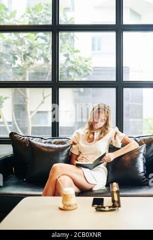 Beautiful young blond woman sitting on leather sofa, drinking sweet refreshing cocktail and writing journal Stock Photo