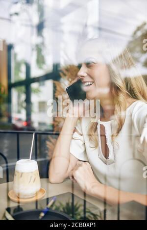 Positive young pretty woman sitting at cafe table and talking on phone with friend Stock Photo