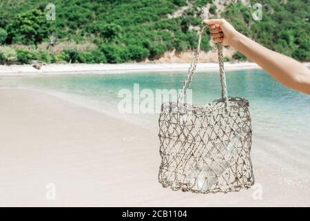 Woman holding straw wicker reusable bag with glass bottle for water. Plastic free and zero waste lifestyle. White sand beach with clear turquoise water on background. Stock Photo