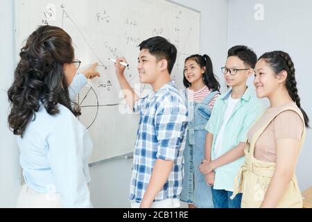 Science teacher explaining theorem to group of students and asking schoolboy to writing equation on whiteboard Stock Photo