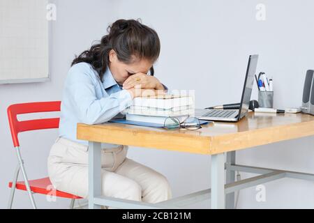 Exhausted teacher leaning on stack on students books on her desk to rest after long stressful day Stock Photo