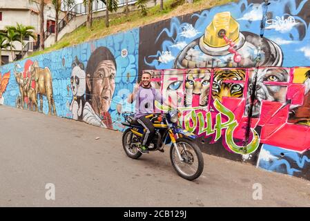 Medellin / Colombia - July 15, 2017: young man making victory sign riding motorcycle in front of graffiti wall in Comuna 13 Stock Photo