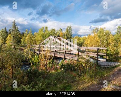 Wooden foot bridge over river stream near Kvikkjokk in Sweden Lapland with birch and spruce tree forest. Early autumn colors, sunlight, blue sky white Stock Photo
