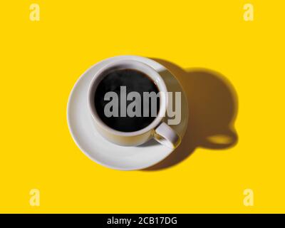 Hot strong black coffee in white elegant cup with saucer over a bright yellow background. Hot morning beverage. Shot with hard light. Top view. Stock Photo