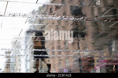Blurry reflection shadow silhouette on wet puddle of a woman under umbrella walking a city street on a rainy day, abstract background Stock Photo