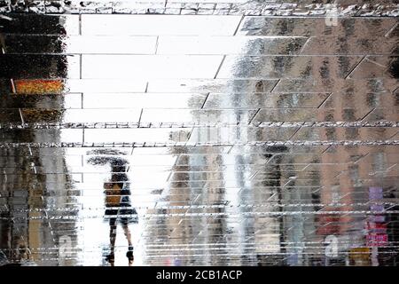 Blurry reflection shadow silhouette on wet puddle of a woman under umbrella walking a city street on a rainy day, abstract background Stock Photo