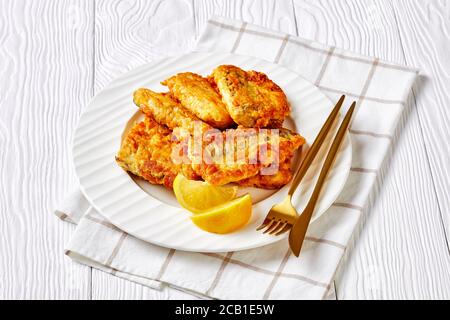 Delicious recipe of battered pan-fried hake fish fillet, served on a white plate with lemon wedges golden cutlery on a white wooden background with na Stock Photo