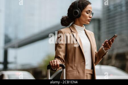 Female executive looking at her mobile phone while standing on a street with luggage. Businesswoman standing on street with luggage listening to music Stock Photo