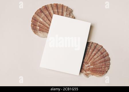 Modern summer stationery still life. Blank vertical greeting card, invitation mock up scene with pink oyster sea shells. Beige table background. Flat Stock Photo