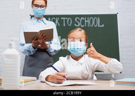 selective focus of schoolgirl in protective mask showing thumb up, and teacher standing near chalkboard with back to school lettering Stock Photo