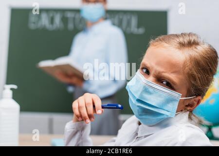 selective focus of frowning schoolgirl in medical mask holding pen and looking at camera, while teacher standing near chalkboard Stock Photo