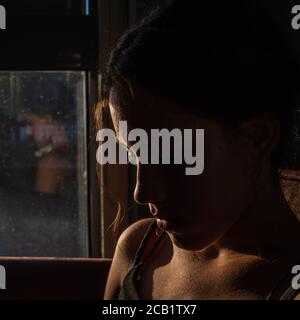 Portrait of beautiful brunette girl in darkness with soft light on her face, pensive silhouette looking down, a reflection on the window.