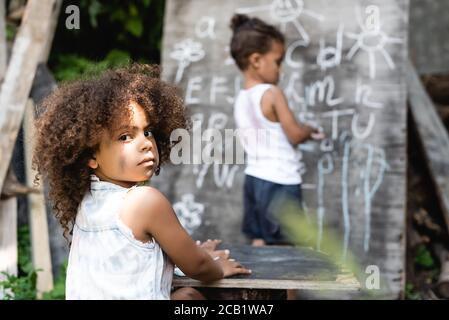 selective focus of curly african american kid sitting near table, boy and chalkboard Stock Photo