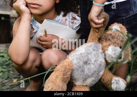 cropped view of poor african american kid holding notebook near child standing with dirty soft toy Stock Photo