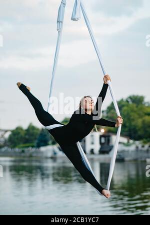 Woman aerialist performs gymnastic split on hanging aerial silk against background of river, sky and trees. Stock Photo