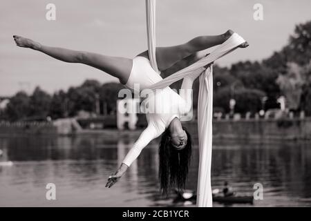 Woman aerialist performs gymnastic split on hanging aerial silk against background of river, sky and trees. Black and white image. Stock Photo